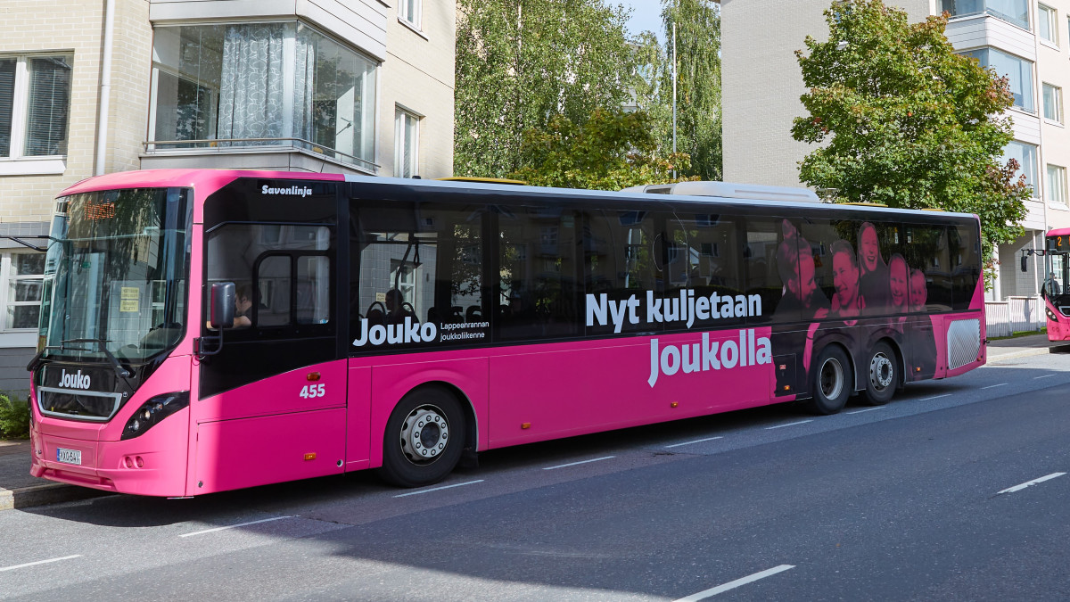 Pinkki linja-auto tiellä. Bussin kyljessä teksti: Kuljetaan Joukolla.