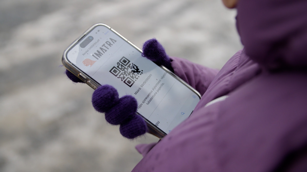 Lilatakkisella henkilöllä kädessään kännykkä, jonka ruudulla Imatran logo ja QR-koodi.