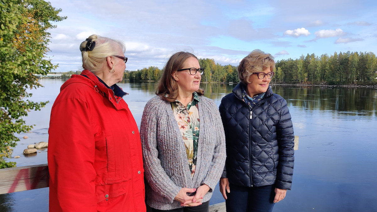 Kolme vanhaa naista seisomassa, taustalla joki.