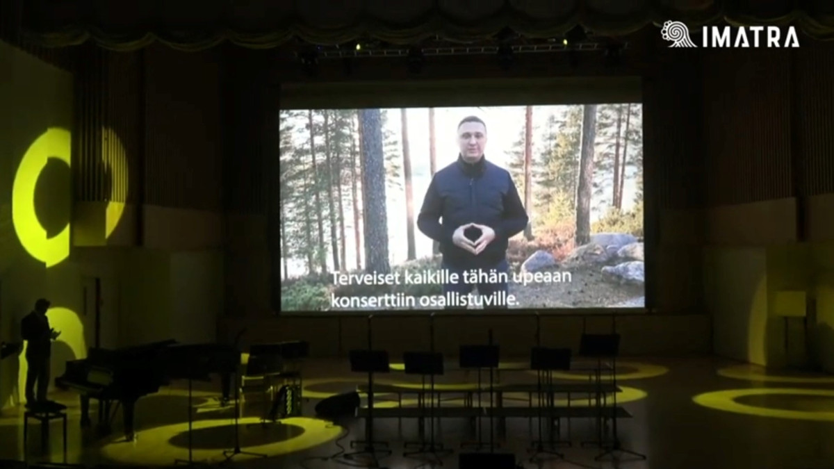 Mies valkokankaalla konserttisalissan näyttämöllä, Näytöllä teksti "Terveiset kaikille tähän upeaan konserttiin osallistuville."