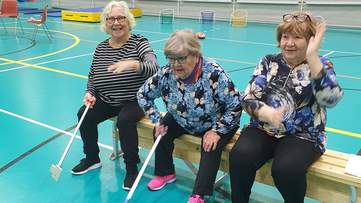Kolme naista istuu hymyssä suin liikuntasalin penkillä.