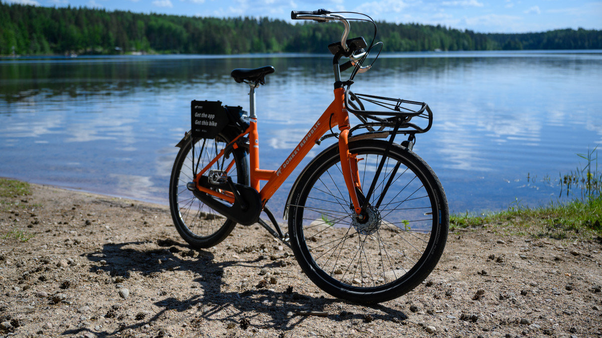 Oranssi kaupunkipyörä hiekkarannalla, taustalla järvi, metsää ja sininen taivas.