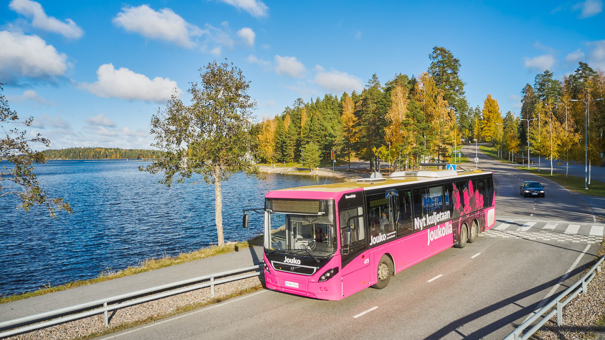 Pinkki bussi ajaa tietä pitkin. Taustalla näkyy järvimaisema. Bussin kyljessä on teksti: Nyt kuljetaan Joukolla.
