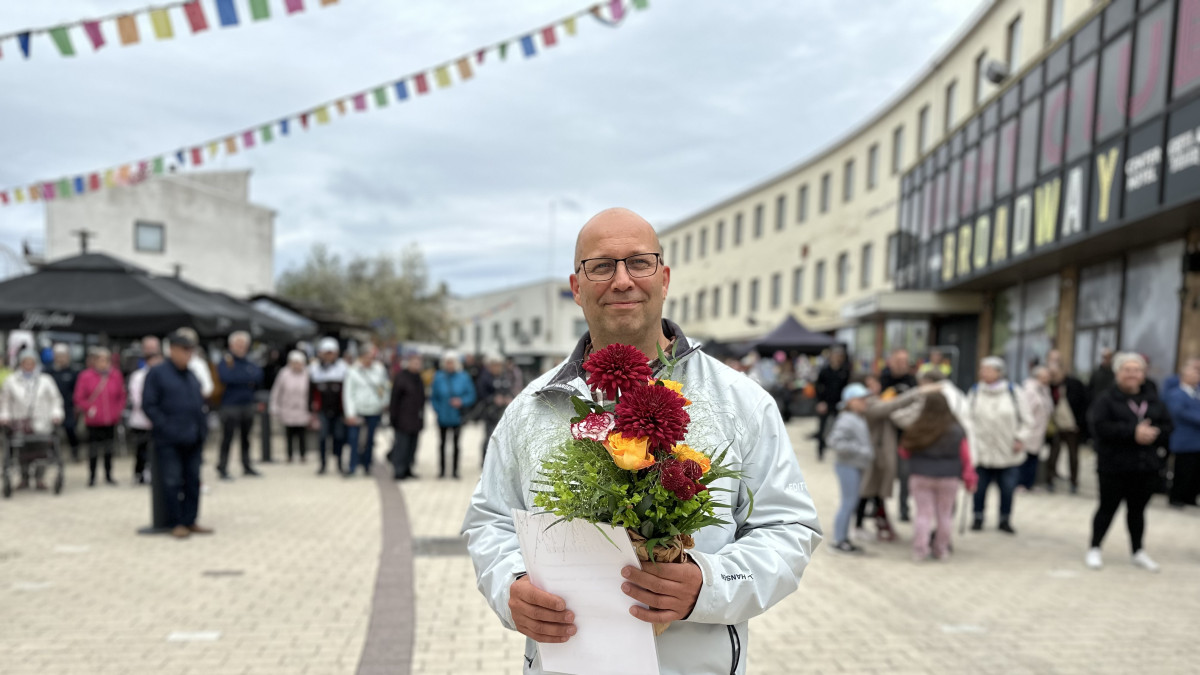 Mies seisoo kävelykadulla kukkakimppu ja kunniakirja käsissään.
