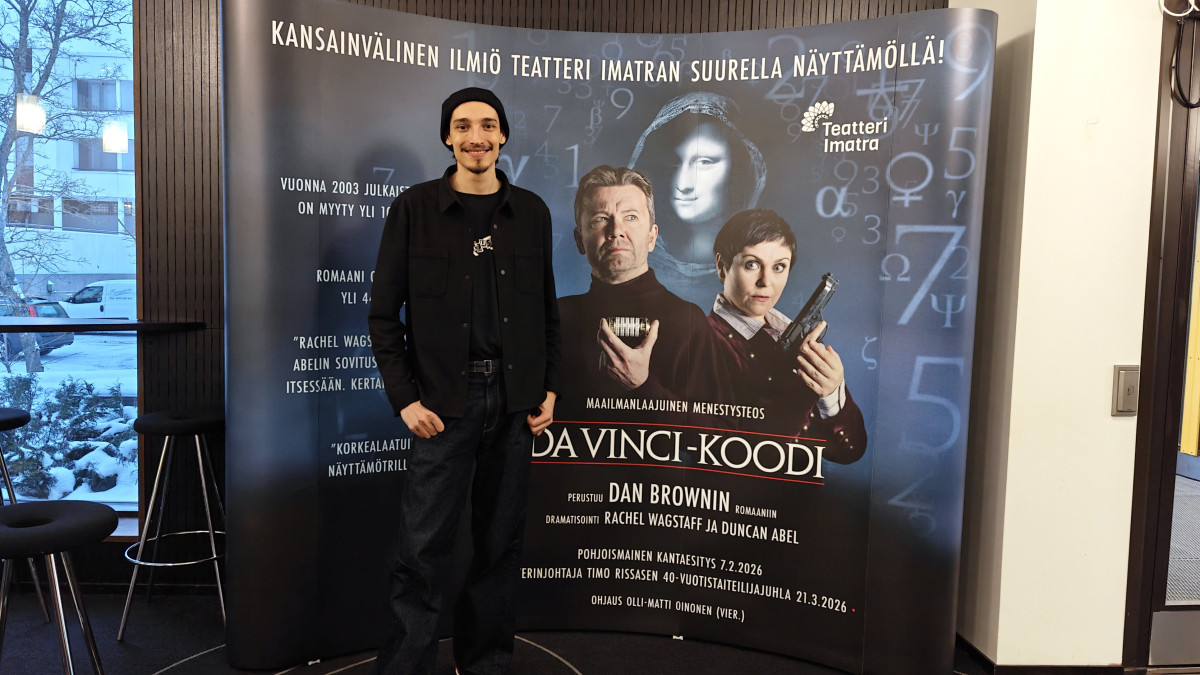 Mies seisoo Da Vinci -koodi mainosseinän edessä ja hymyilee kameralle.