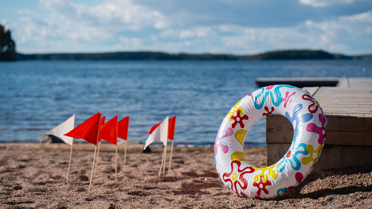 Hiekkaranta, vettä, uimarengas, laituri ja pikkulippuja hiekassa.