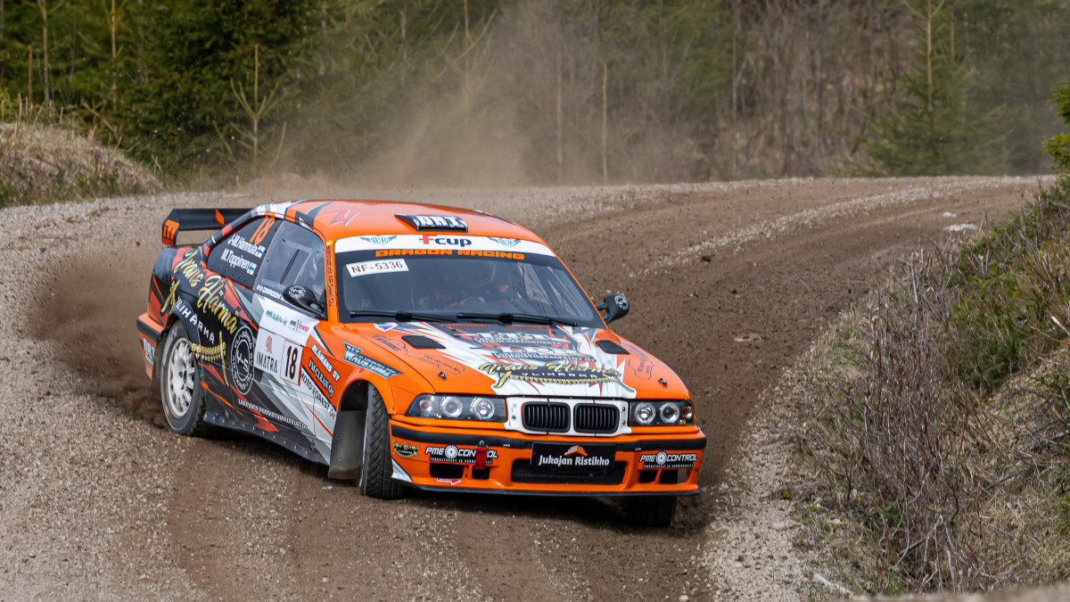 Ornassivalkoinen ralliauto ajaa hiekkatiellä mutkassa.