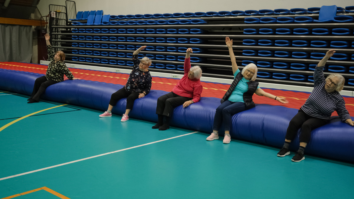 Viisi osallistujaa istuu liikuntasalissa suurella ilmapatjalla ja tekee ohjattua venytys- tai jumppaliikettä kädet ylös ojennettuina. Taustalla näkyvät siniset katsomopenkit.