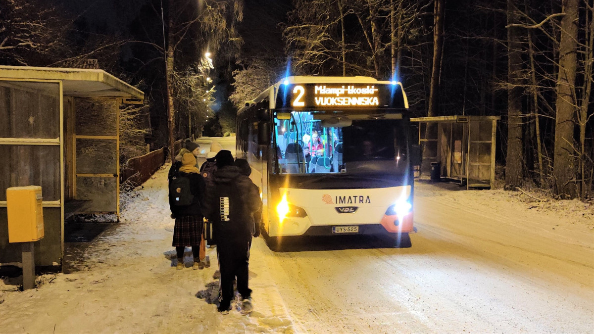 Linja-auto saapumassa pysäkille ja koululaiset odottamassa sitä talvisäässä.