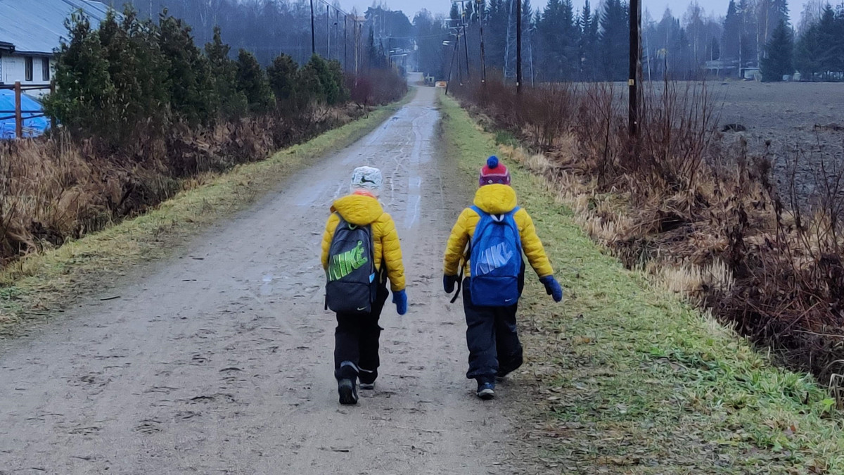 kaksi pientä poikaa samanvärisissä takeissa reput selässä selkäpuolelta kuvattuna kävelemässä suoraa hiekkatietä peltojen halki.