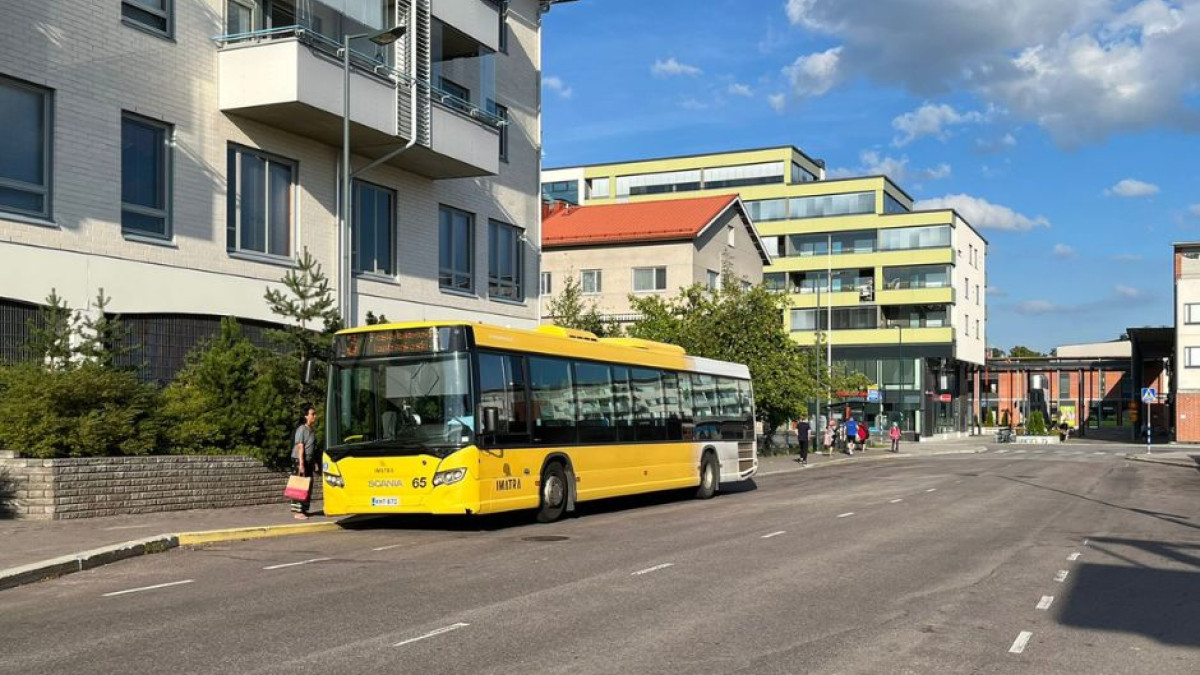 Bussi bussipysäkillä. Bussin ympärillä on ihmisiä jalkakäytävällä ja pysäkillä.