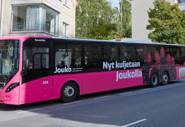 Pinkki linja-auto tiellä. Bussin kyljessä teksti: Kuljetaan Joukolla.