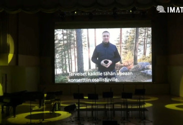 Mies valkokankaalla konserttisalissan näyttämöllä, Näytöllä teksti "Terveiset kaikille tähän upeaan konserttiin osallistuville."
