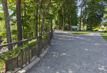 Hiekkaisen puistotien reunassa takorautainen aita ja vihreää puustoa kesäauringon paisteessa