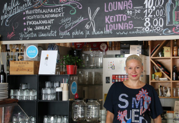 Yrittäjä seisoo ravintolansa tiskin edessä, ylhäällä liitutauluun kirjoitettuna ravintolassa olevat tarjottavat.
