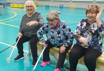 Kolme naista istuu hymyssä suin liikuntasalin penkillä.