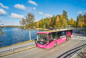 Pinkki bussi ajaa tietä pitkin. Taustalla näkyy järvimaisema. Bussin kyljessä on teksti: Nyt kuljetaan Joukolla.