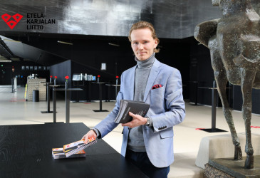 Henkilö (Miika Asikainen) pitelee käsissään teatterin esitteitä. Taustalla näkyy Lappeenrannan kaupunginteatterin lämpiö, jossa muun muassa pöytiä. Kuvan päällä Etelä-Karjalan liiton logo.