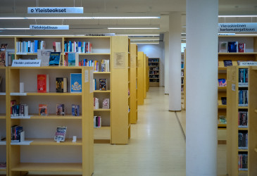 Kirjaston kirjahyllyjä, joiden välissä menee käytävä. Käytävällä valkoisia pylväitä peräkkäin.