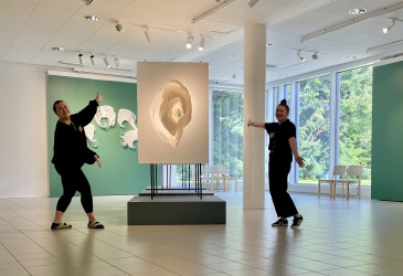 Kaksi henkilöä poseeraa taidemuseossa suuren taideteoksen vieressä. Teos on asetettu jalustalle ja siinä on pyörteisiä, vaaleita muotoja. Tilassa on suuret ikkunat, joista näkyy vehreä maisema. Seinillä on lisää taideteoksia ja ikkunoiden lähellä on tuoleja.