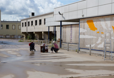 Henkilö työskentelee ulkotilassa vanhan liiketilan edustalla, jossa vanerilla peitettyyn seinään on maalattu valkoisia ja oransseja muotoja. Taustalla näkyy rakennuksia.