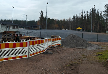 Työmaa-aitojen ja hiekka-alueen takana asfaltoitu kenttä.