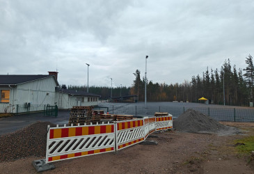 Asfaltoitu alue, jonka edessä on rakennusaidat. Alueen vasemmalla puolella on vaalea huoltorakennus ja takana metsää.