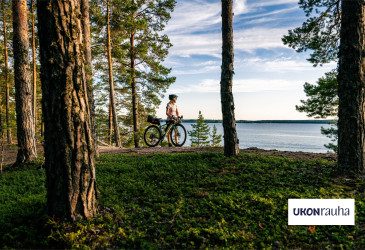 Pyöräilijä seisoo pyörän vieressä ja katsoo järvelle. Ympärillä on metsää. Kuvan päällä Ukonrauha-logo.