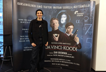 Mies seisoo Da Vinci -koodi mainosseinän edessä ja hymyilee kameralle.