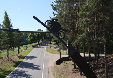 Kävelysillalta kuvattu näkymä metsän reunustamalle maantielle. Etualalla moderni katuvalopylväs, jonka varressa metallinen moottoripyöräkoriste.