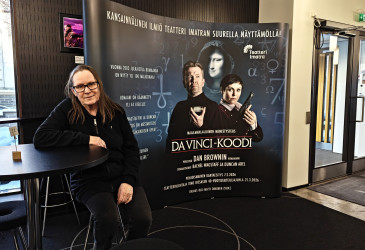 Näyttelijä istuu tuolilla ja nojaa pöytään. Taustalla näkyy ikkunat sekä Da Vinci -koodin mainosseinä.