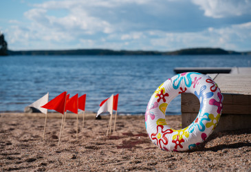 Hiekkaranta, vettä, uimarengas, laituri ja pikkulippuja hiekassa.