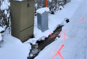 Ojassa vettä sähkökaapin vieressä, lumessa oranssilla maalilla merkintöjä.