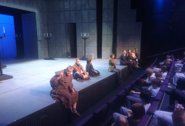 Teatterin näyttämö, jossa useita esiintyjiä istuu lattialla ja näyttämön reunalla. Taustalla on harmaa, kivimäinen lavastus ja kynttiläjalustoja. Etualalla näkyy katsomossa istuva yleisö, joka seuraa esitystä.