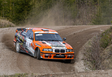Ornassivalkoinen ralliauto ajaa hiekkatiellä mutkassa.