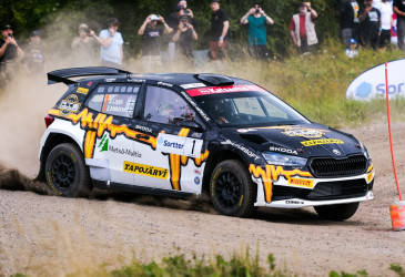 Ralliauto mutkassa, sora pöllyää, yleisöä taustalla.