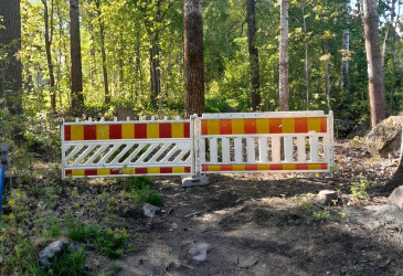 Metsäisessä maastossa kulkeva polku on suljettu kahdella vierekkäisellä valkoisella työmaa-aidalla, joissa on punakeltaiset heijastinraidat. Taustalla kasvaa tiheää vihreälehtistä puustoa.