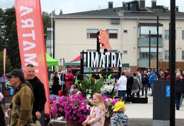 Kuvassa Imatran Napinkulman aukiolla kaupunkilaisia, kesäkukkia ja kaupungin tunnuksia.