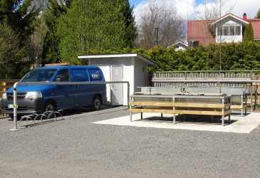 Ulkoilmassa sijaitseva matonpesupaikka, jossa on useita pesupöytiä ja vedenottohanat. Vieressä on pieni valkoinen huoltorakennus ja sininen pakettiauto sorapihalla. Taustalla näkyy puuaita, puita sekä punakattoisia pientaloja aurinkoisessa säässä.