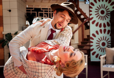 Kaksi näyttelijää teatterilavalla tanssiasennossa: hatullinen henkilö pitelee toista henkilöä taaksepäin taivutettuna. Molemmilla on vaaleat, 1900-luvun alun tyyliä muistuttavat asut, ja taustalla näkyy sisätilaa esittävä lavastus portaikkoineen ja kuvioituine seinäpintoineen kirkkaassa valaistuksessa.