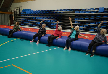 Viisi osallistujaa istuu liikuntasalissa suurella ilmapatjalla ja tekee ohjattua venytys- tai jumppaliikettä kädet ylös ojennettuina. Taustalla näkyvät siniset katsomopenkit.