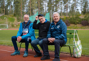 Kolme iäkästä miestä istuu penkillä urheilukentän laidalla.