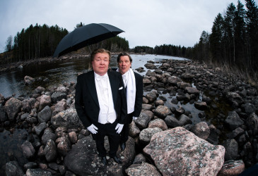 Kaksi frakkiin pukeutunutta henkilöä seisoo kivikkoisella joenrannalla. Toisen yläpuolella on avoin musta sateenvarjo. Taustalla näkyy virtaava joki, runsaasti kiviä sekä metsää harmaan pilvisen taivaan alla.