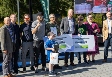 Ulkoilmatilaisuudessa ryhmä ihmisiä poseeraa ympäristöpalkintojen jakotilaisuudessa. Kuvassa pidetään esillä Etelä-Karjalan ympäristöpalkinnon pääpalkintoa (10 000 euroa) ja elämäntyöpalkintoa (1 000 euroa) sekä kunniakirjaa. Etualalla lapsi pitelee palkintolahjakassia.