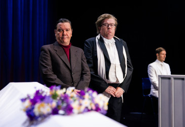 Teatterin näyttämöllä kaksi tummaan juhla-asuun pukeutunutta esiintyjää seisoo valokeilassa valkoisen arkun vieressä. Arkun päällä on violeteista ja vaaleista kukista koottu kukka-asetelma. Taustalla näkyy sinisellä valaistu verho sekä valkoiseen asuun pukeutunut henkilö. Näyttämön valaistus korostaa muistotilaisuuden tai hautajaiskohtauksen tunnelmaa.