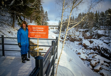 Mies karvahatussa seisoo Imatrankosken rannalla.  Kyltissä teksti: kaupunginjohtajaksi kansallisihmeeseen.