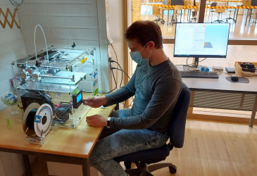 Mieshenkilö istuu 3D-tulostimen ääressä, taustalla on tietokone.
