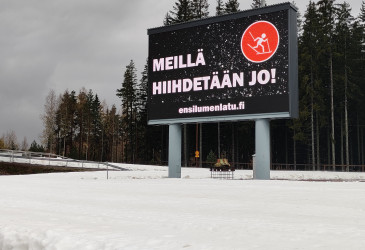 Ensilumenladun kyltti, jossa lukee meillä hiihdetään jo.