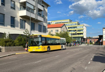 Bussi bussipysäkillä. Bussin ympärillä on ihmisiä jalkakäytävällä ja pysäkillä.