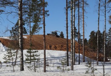Osittain purulla peitetty lumikasa metsikön takana, päällä kauhakuormaaja.